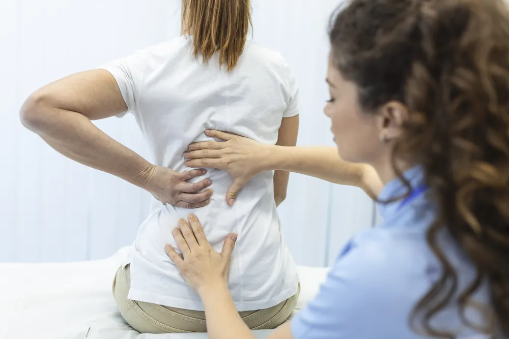 Cimentoplastie : une soignante examine le dos d’une patiente assise qui se tient le bas du dos, douloureux.