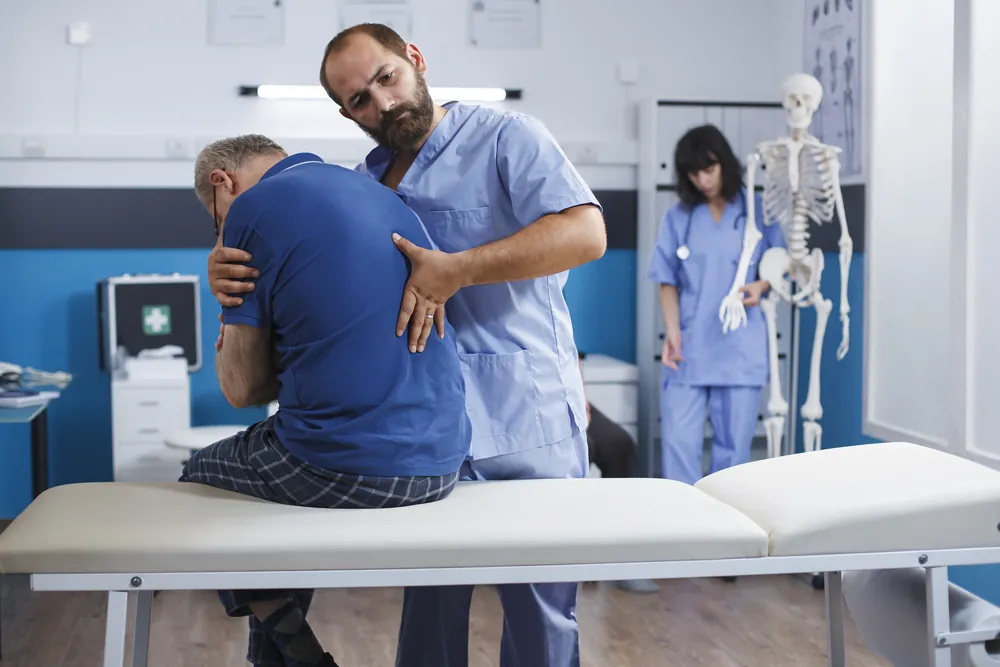 Cruralgie : un soignant examine le bas du dos d’un patient assis sur une table d’examen, penché en avant sous l’effet de la douleur.