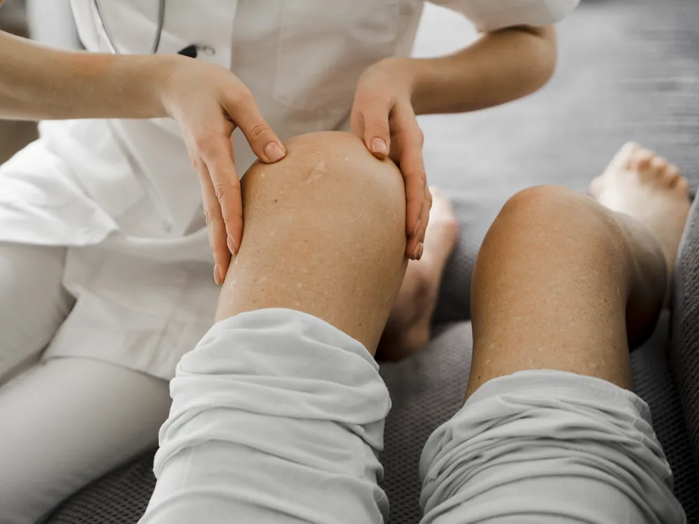 Gonalgie : une médecin en blouse palpe le genou fléchi d’un patient assis pour localiser la douleur.
