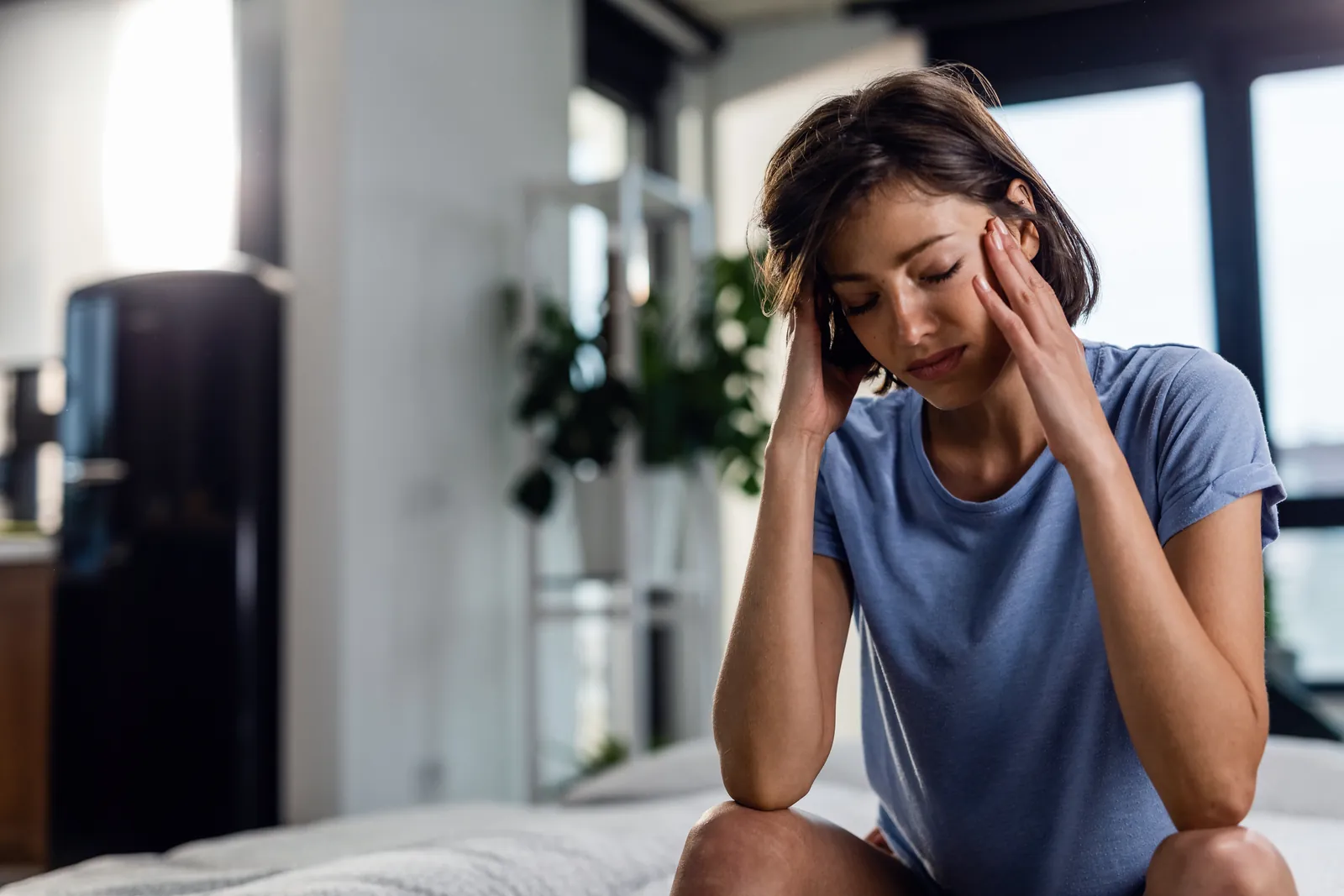 Névralgie : une femme assise sur son lit, les yeux fermés, se tient la tête des deux mains sous l’effet de la douleur.