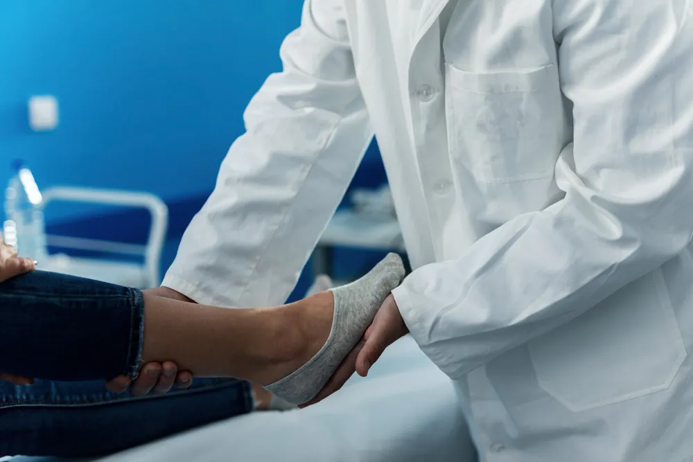 Un médecin en blouse blanche examine le pied d’une patiente allongée, en tenant l’avant-pied dans sa main.