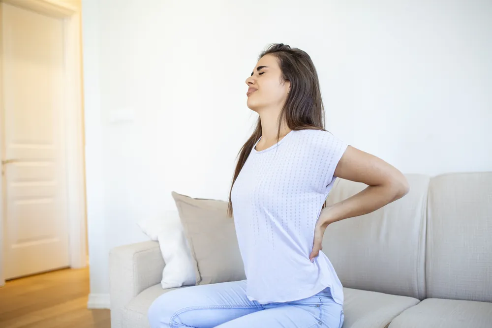Rhizolyse : une femme assise sur un canapé se masse le bas du dos, le visage crispé.