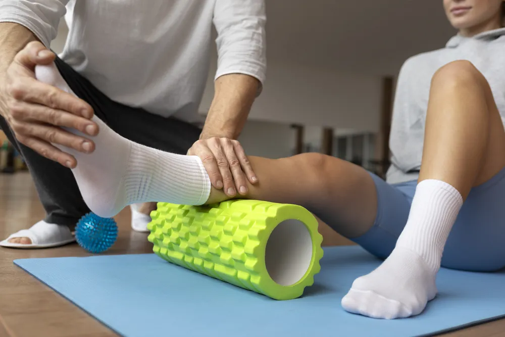 Traitement du névrome de Morton : un thérapeute accompagne une patiente lors d’exercices du pied et de la jambe, le mollet posé sur un rouleau de massage.