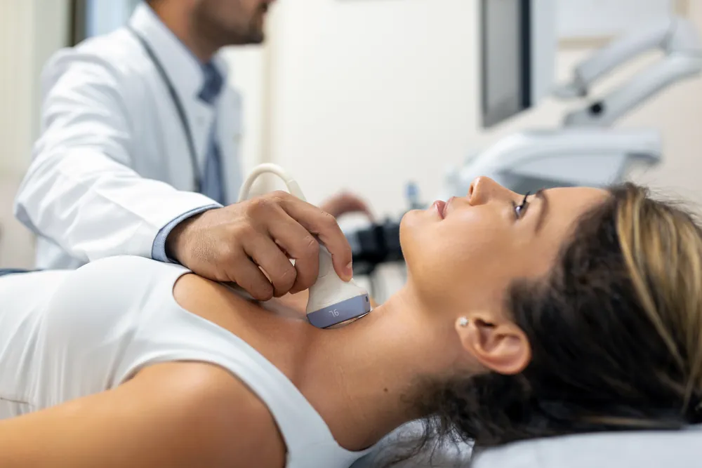 Cytoponction thyroïdienne guidée par échographie : un médecin pose la sonde sur le cou d’une patiente allongée.